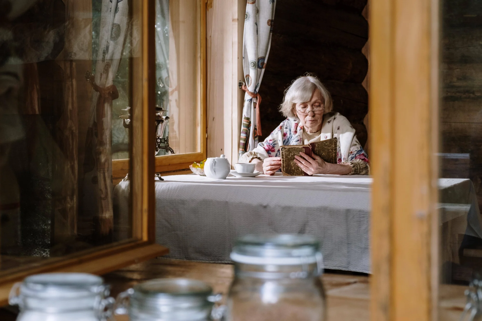 Mujer mayor con cabello blanco y gafas lee un libro antiguo sentada junto a una ventana, con una tetera y taza sobre la mesa.