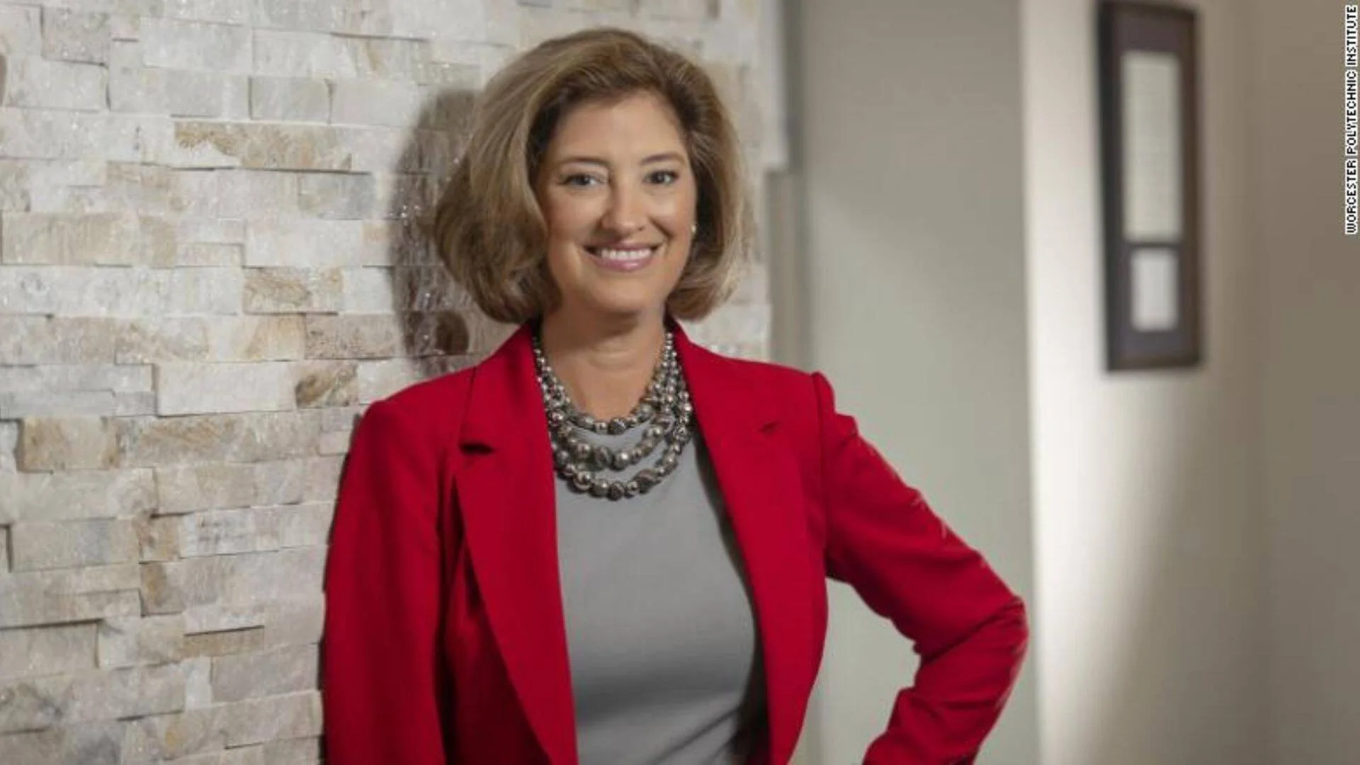 Mujer sonriente con blazer rojo y collar plateado apoyada en una pared de piedra, en foto oficial del Worcester Polytechnic Institute.