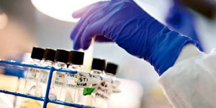 Técnico con guante azul toma un vial de una gradilla con muestras etiquetadas en un laboratorio.