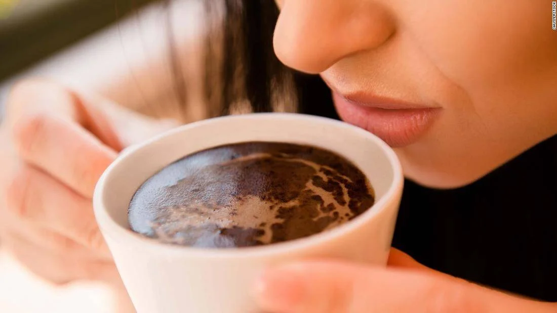 Una mujer acerca su rostro a una taza de café negro humeante que sostiene con ambas manos.
