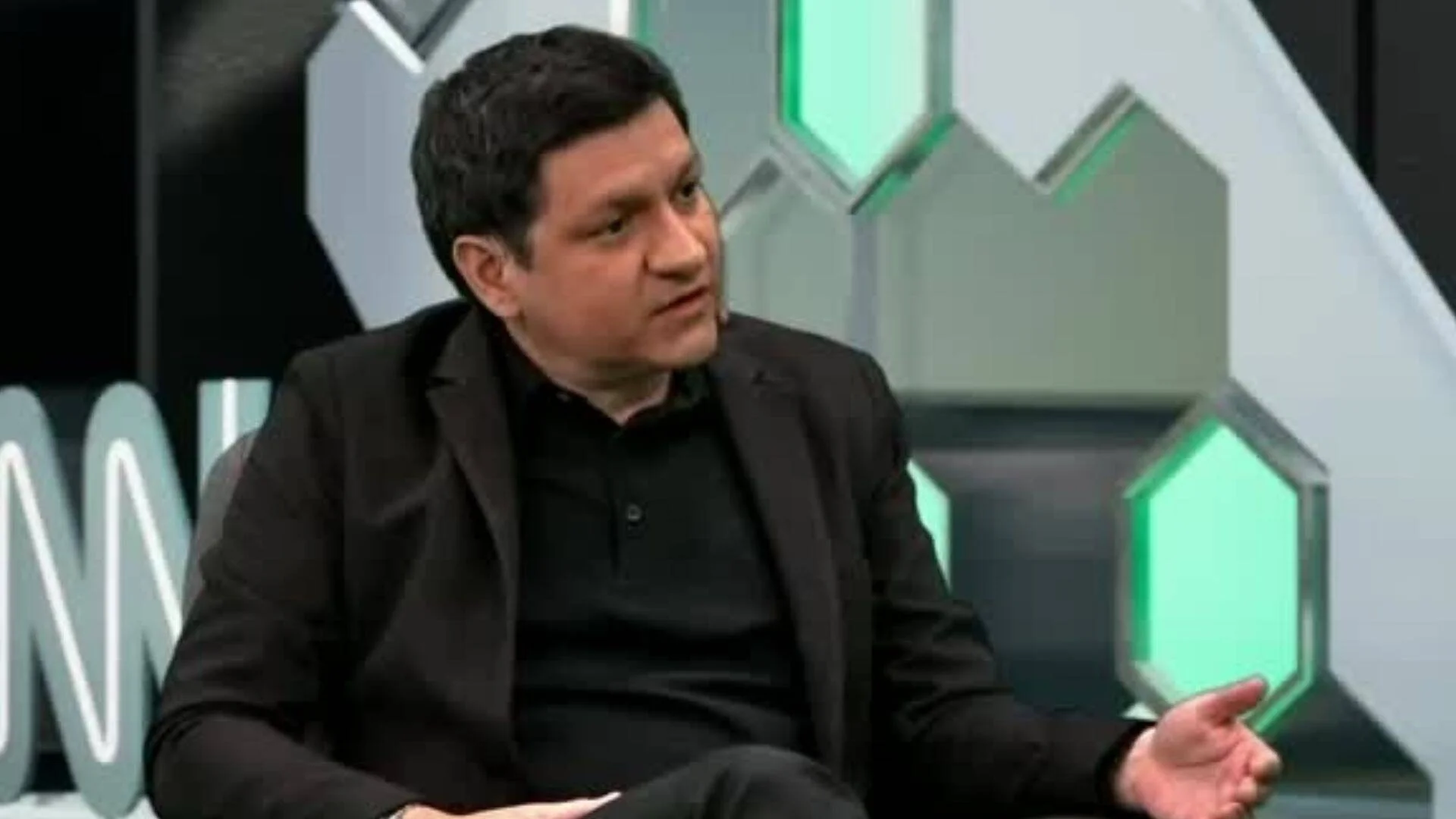 Hombre vestido de negro habla gesticulando en un estudio de televisión con decorado geométrico verde y gris.
