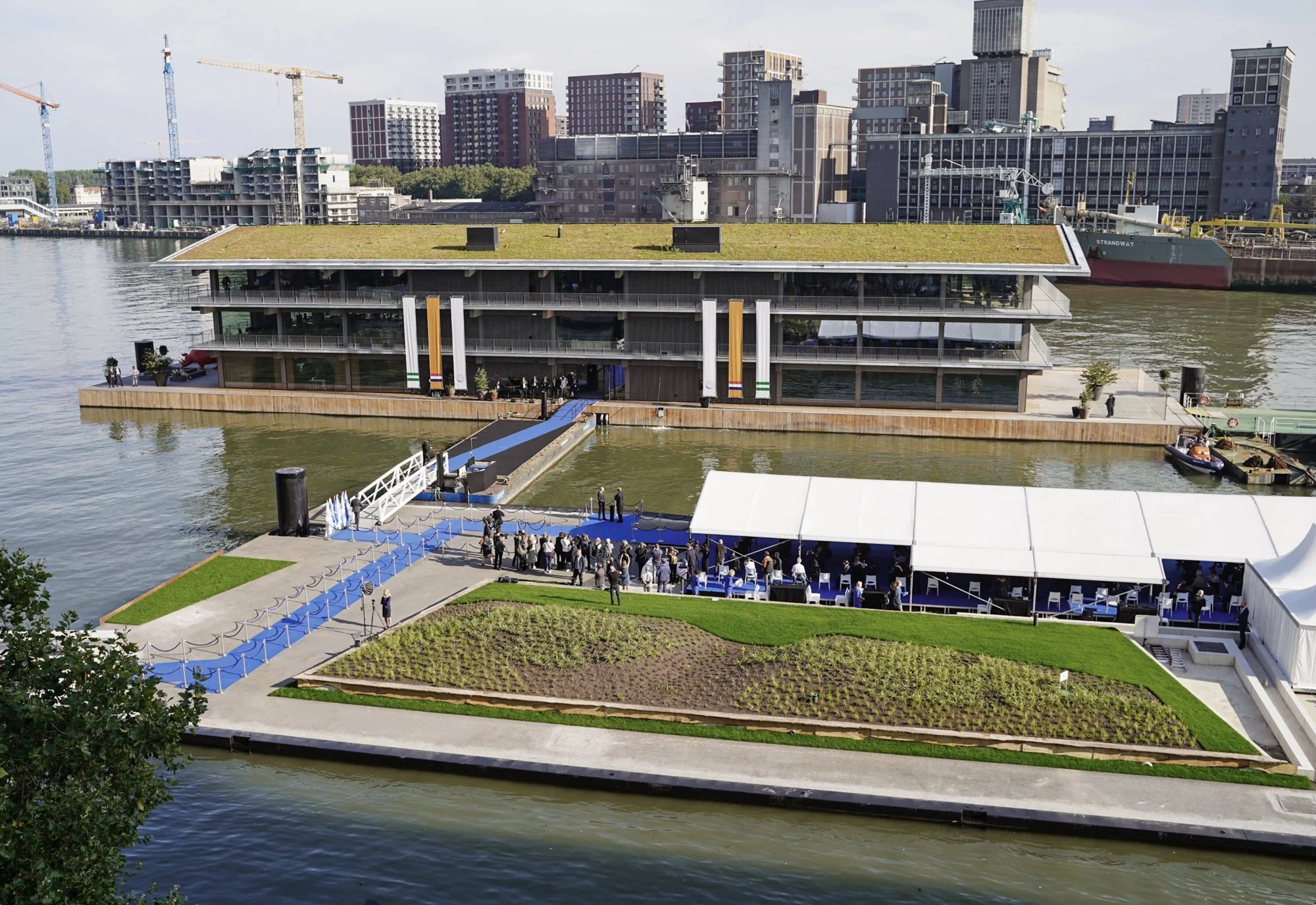 Edificio moderno flotante con techo ajardinado sobre el río, rodeado de invitados en un evento de inauguración con alfombra azul.