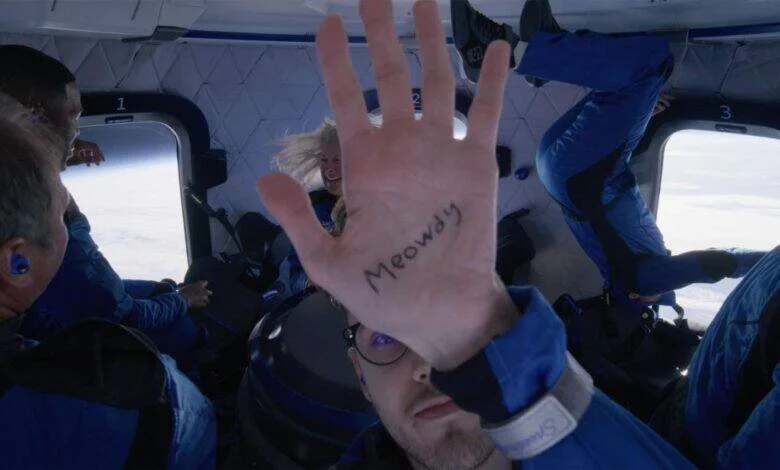 Pasajero en cápsula espacial en ingravidez muestra la palma de su mano con la palabra 'Meowdy' escrita, mientras otros tripulantes flotan al fondo.