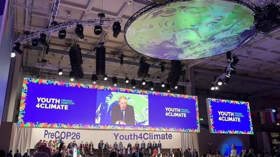 Sala del evento Youth4Climate PreCOP26 con pantallas gigantes que muestran la transmisión de un orador en el escenario.