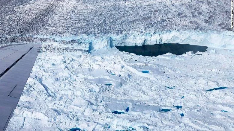 Vista aérea desde avión de un glaciar fracturado con una laguna de agua oscura entre bloques de hielo y acantilado glaciar.