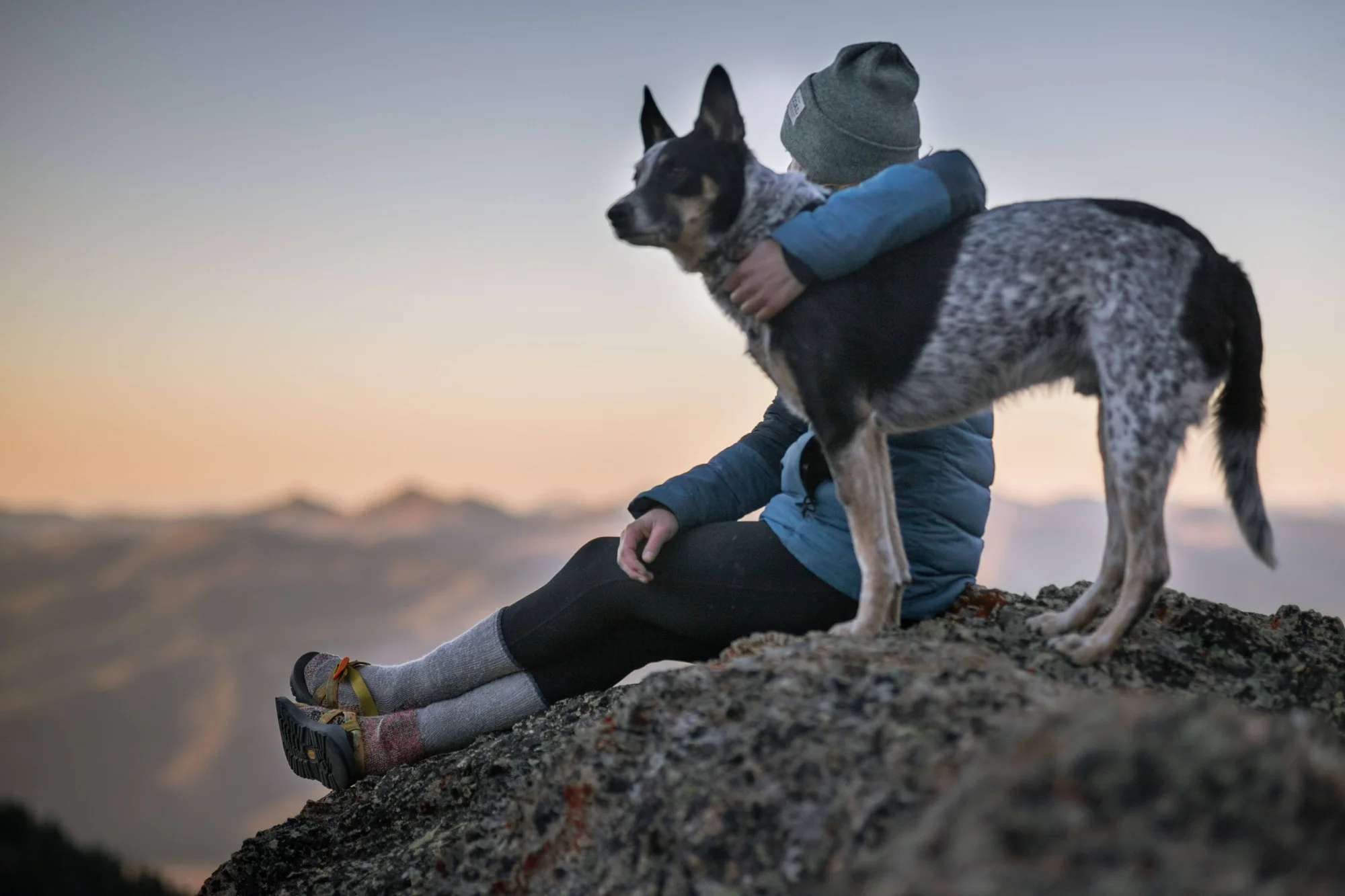 Una persona sentada en una roca en la cima de una montaña abraza a su perro mientras contemplan el horizonte al atardecer.