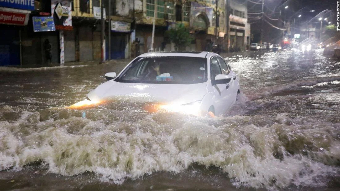 Un automóvil blanco avanza con dificultad por una calle completamente inundada durante la noche, levantando grandes olas de agua.