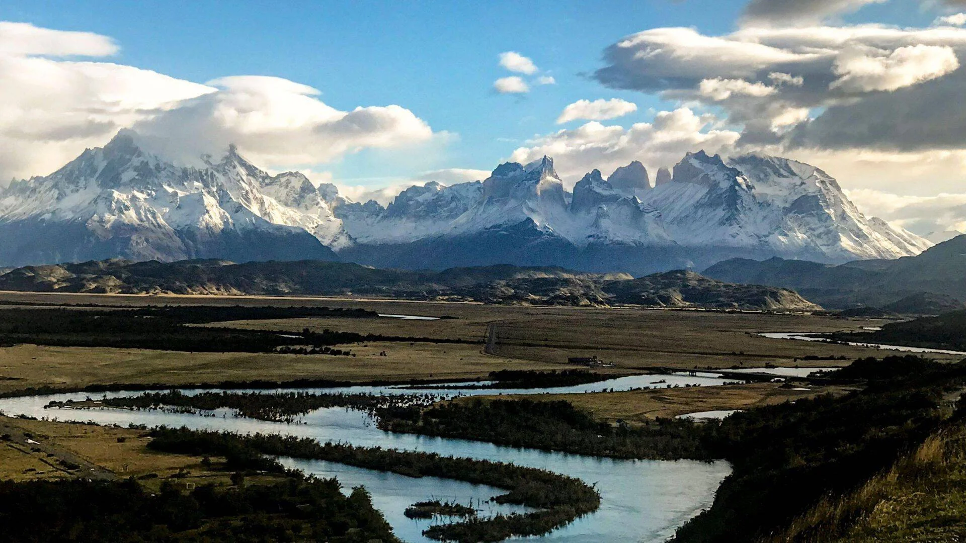 Río de curso sinuoso atraviesa una llanura patagónica con macizo montañoso nevado y torres rocosas al fondo bajo cielo nublado.