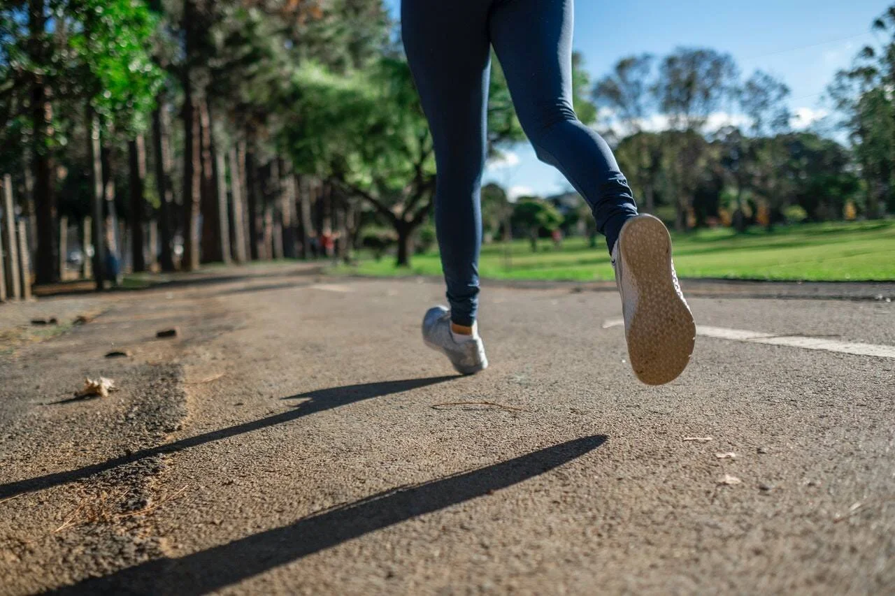 Vista a ras del suelo de las piernas de una persona corriendo con zapatillas blancas y calzas azules en un parque arbolado.