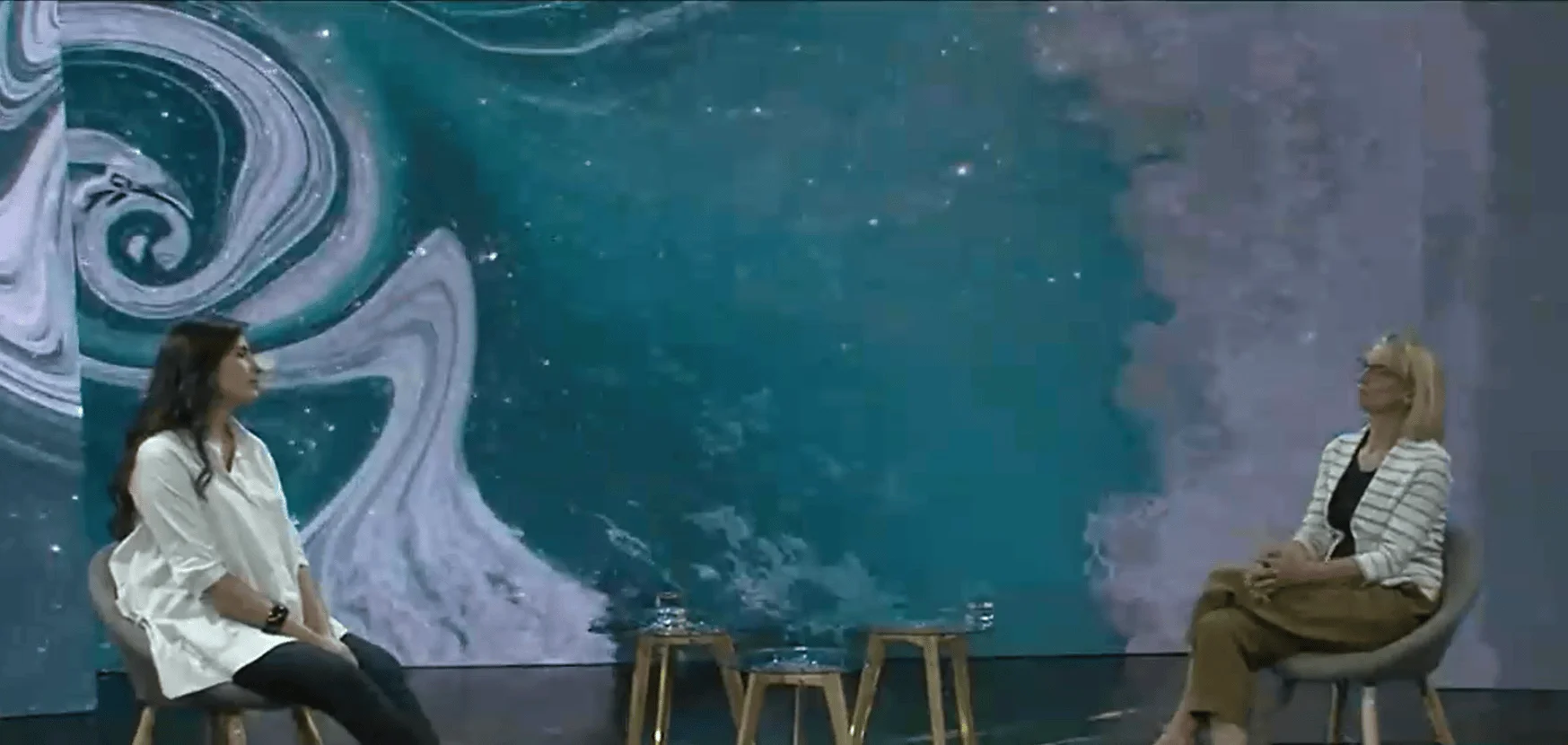 Dos mujeres sentadas frente a frente en un escenario con fondo de arte digital en tonos turquesa y gris.