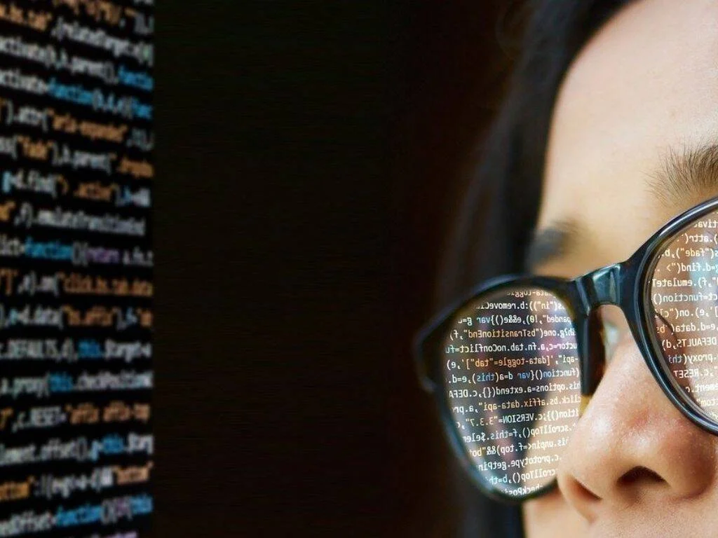 Persona con anteojos mirando una pantalla; el código de programación se refleja en los cristales de sus lentes.