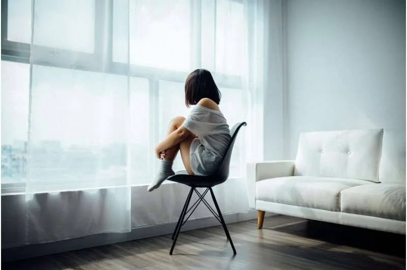 Mujer joven sentada de espaldas en una silla, abrazando sus rodillas y mirando por la ventana en actitud melancólica.