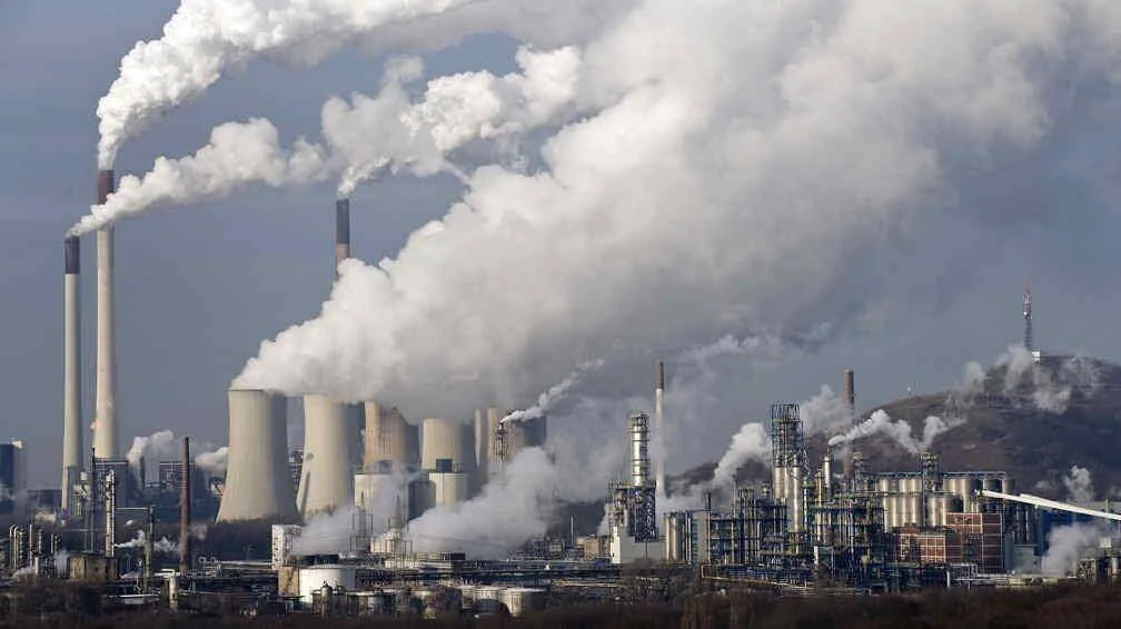 Gran complejo industrial con múltiples chimeneas y torres de refrigeración expulsando densas columnas de humo y vapor al cielo.