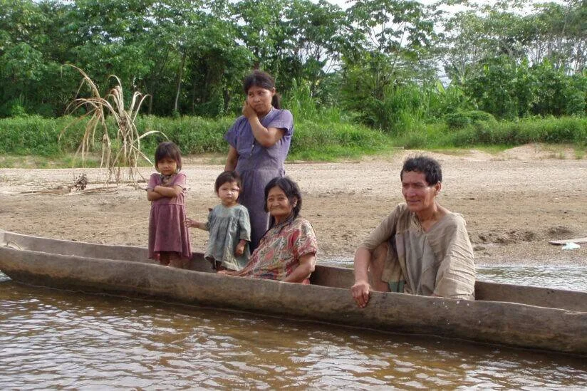 Familia indígena compuesta por adultos mayores, una joven y dos niñas pequeñas en una canoa de madera a orillas de un río amazónico.