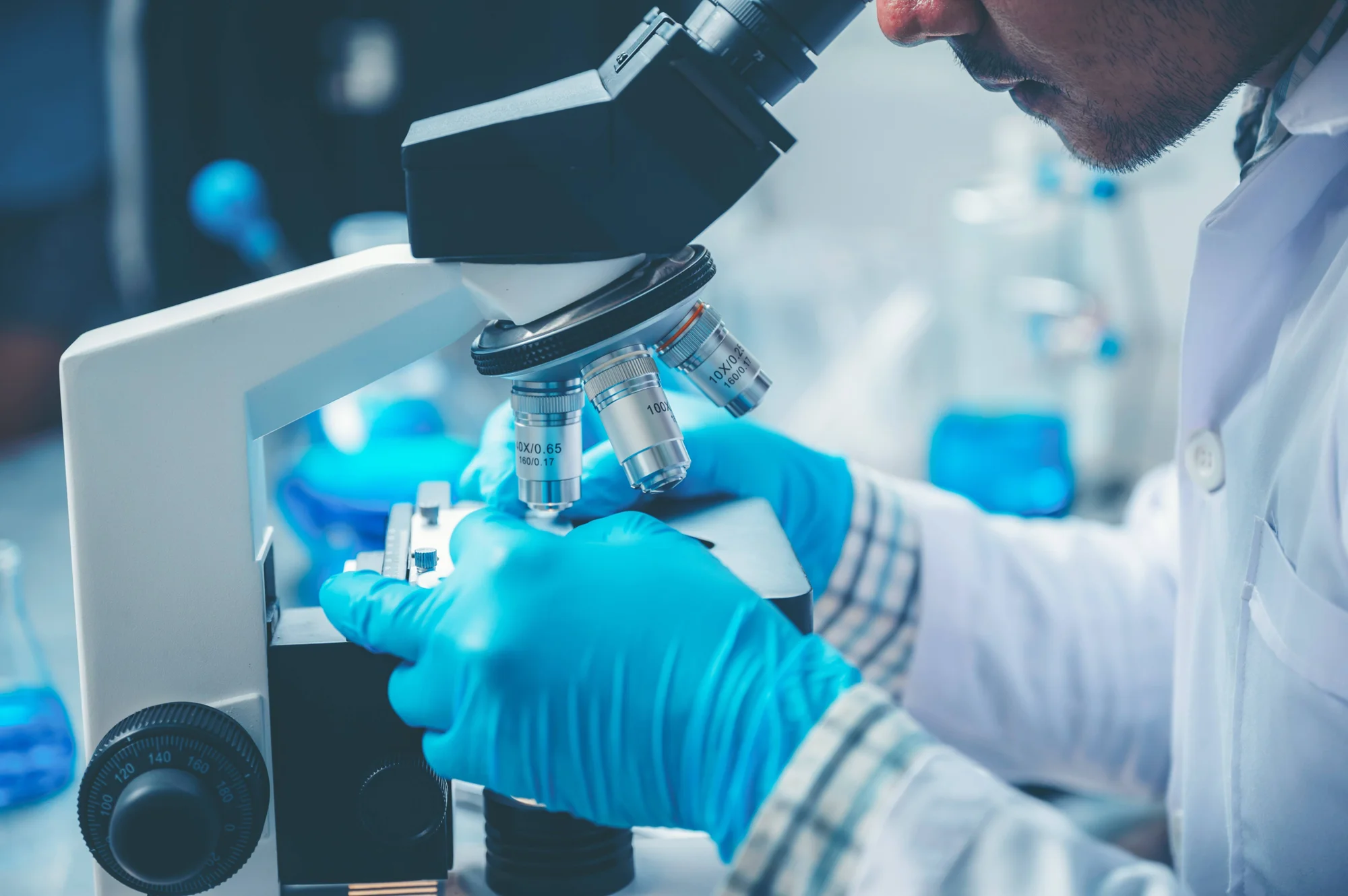 Investigador con guantes azules y bata blanca observa una muestra a través de un microscopio en un laboratorio.