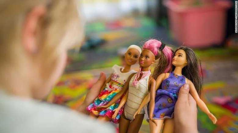 Una niña sostiene tres muñecas Barbie de distintas apariencias y atuendos coloridos en una habitación de juegos.