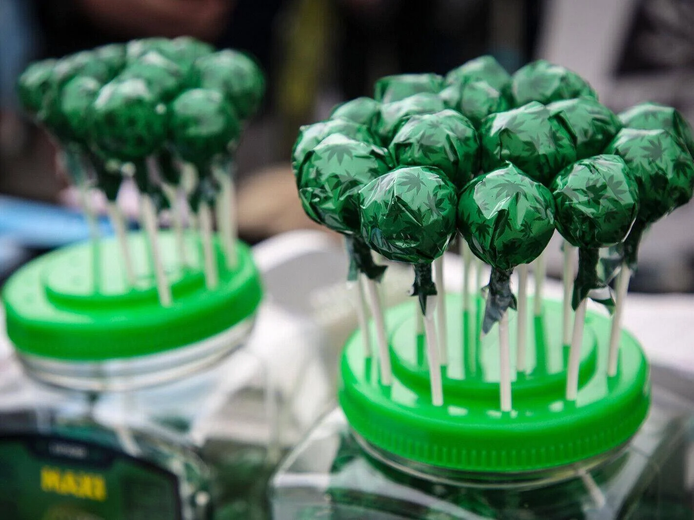 Chupetines envueltos en papel verde con estampado de hojas de cannabis, exhibidos en frascos sobre una superficie de vidrio.