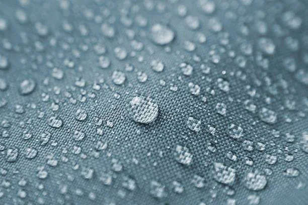 Gotas de agua repelidas sobre la superficie de una tela impermeable de color azul grisáceo, mostrando el efecto hidrófobo del material.