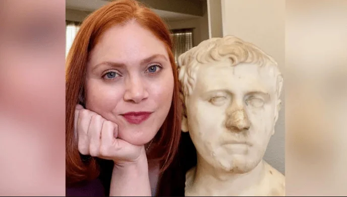 Mujer de cabello rojo posa junto a un busto de mármol de estilo romano antiguo.
