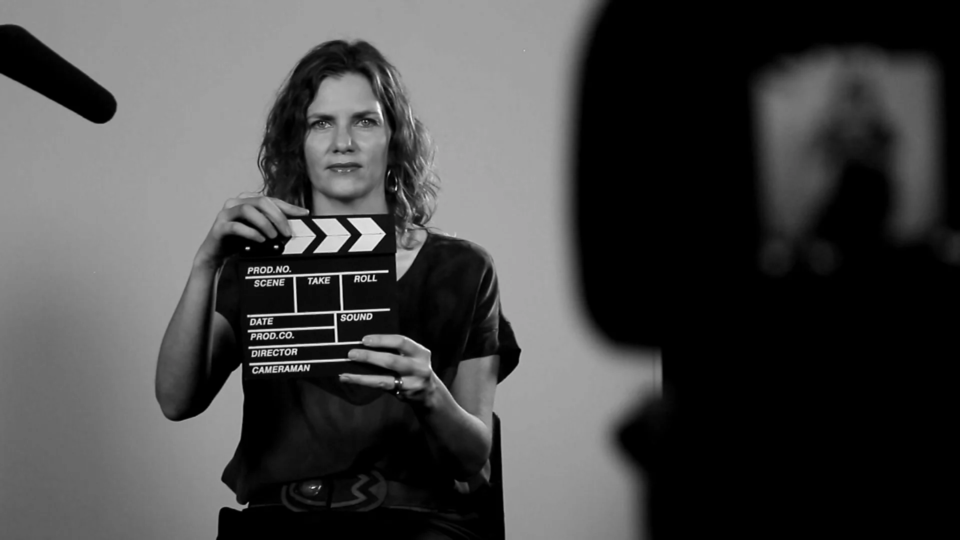 Mujer frente a cámara sostiene una claqueta de cine en un set de filmación, fotografía en blanco y negro.