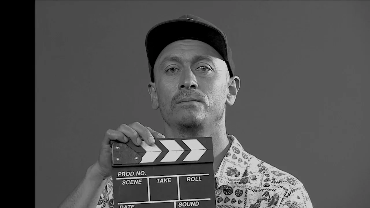 Hombre con gorra sosteniendo una claqueta de filmación frente a cámara, en retrato blanco y negro.