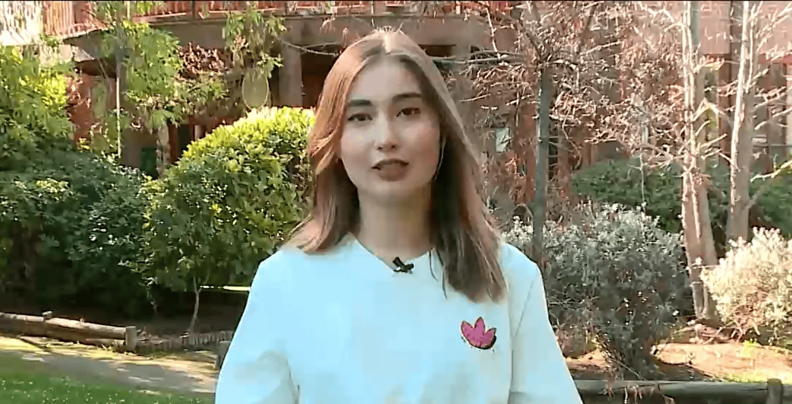 Mujer joven con remera blanca y micrófono de solapa habla a cámara en un parque arbolado.