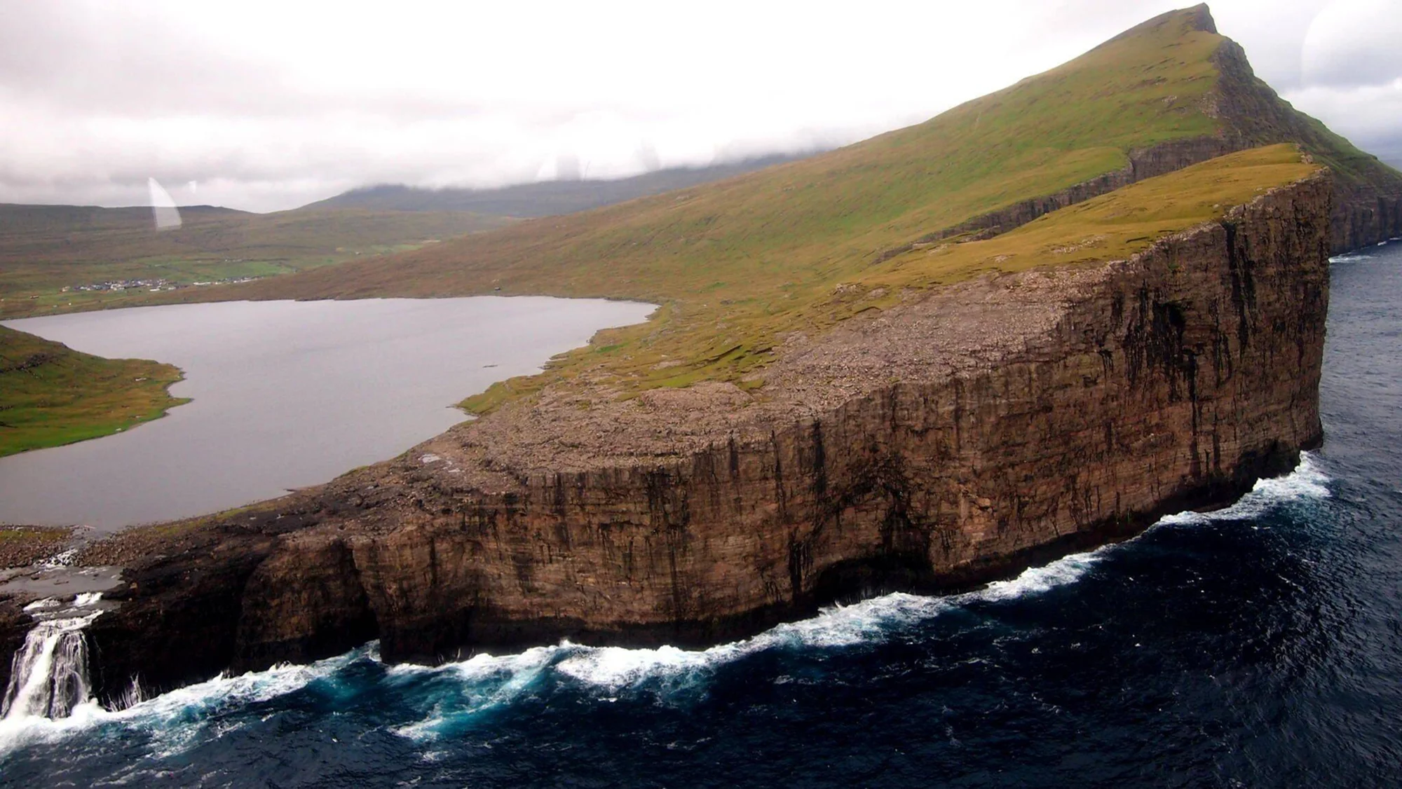Vista aérea de un lago sobre un acantilado rocoso del que cae una cascada directamente al océano, rodeado de colinas verdes bajo cielo nublado.