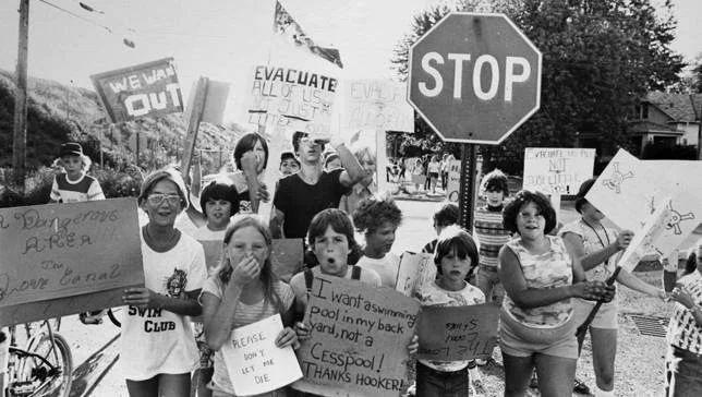 Niños y jóvenes marchan con carteles exigiendo evacuación y denunciando contaminación en Love Canal, junto a una señal de Stop.