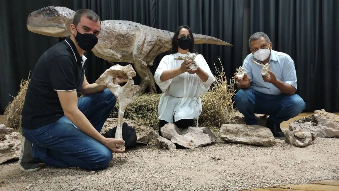 Tres personas con barbijo sostienen fósiles frente a una réplica de dinosaurio en una exposición paleontológica.