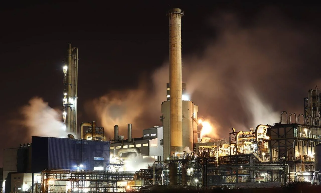 Complejo industrial iluminado de noche con chimeneas expulsando gases y vapor al cielo oscuro.