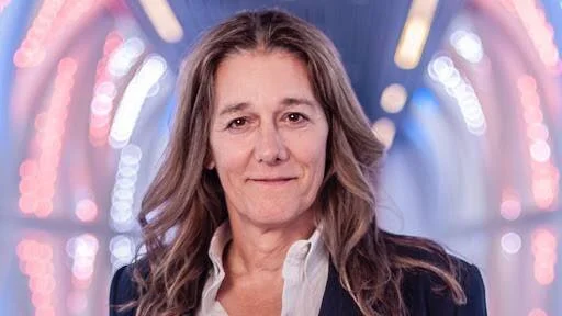 Mujer de mediana edad con cabello largo castaño claro, vestida con blazer oscuro, posando ante un fondo de luces desenfocadas.