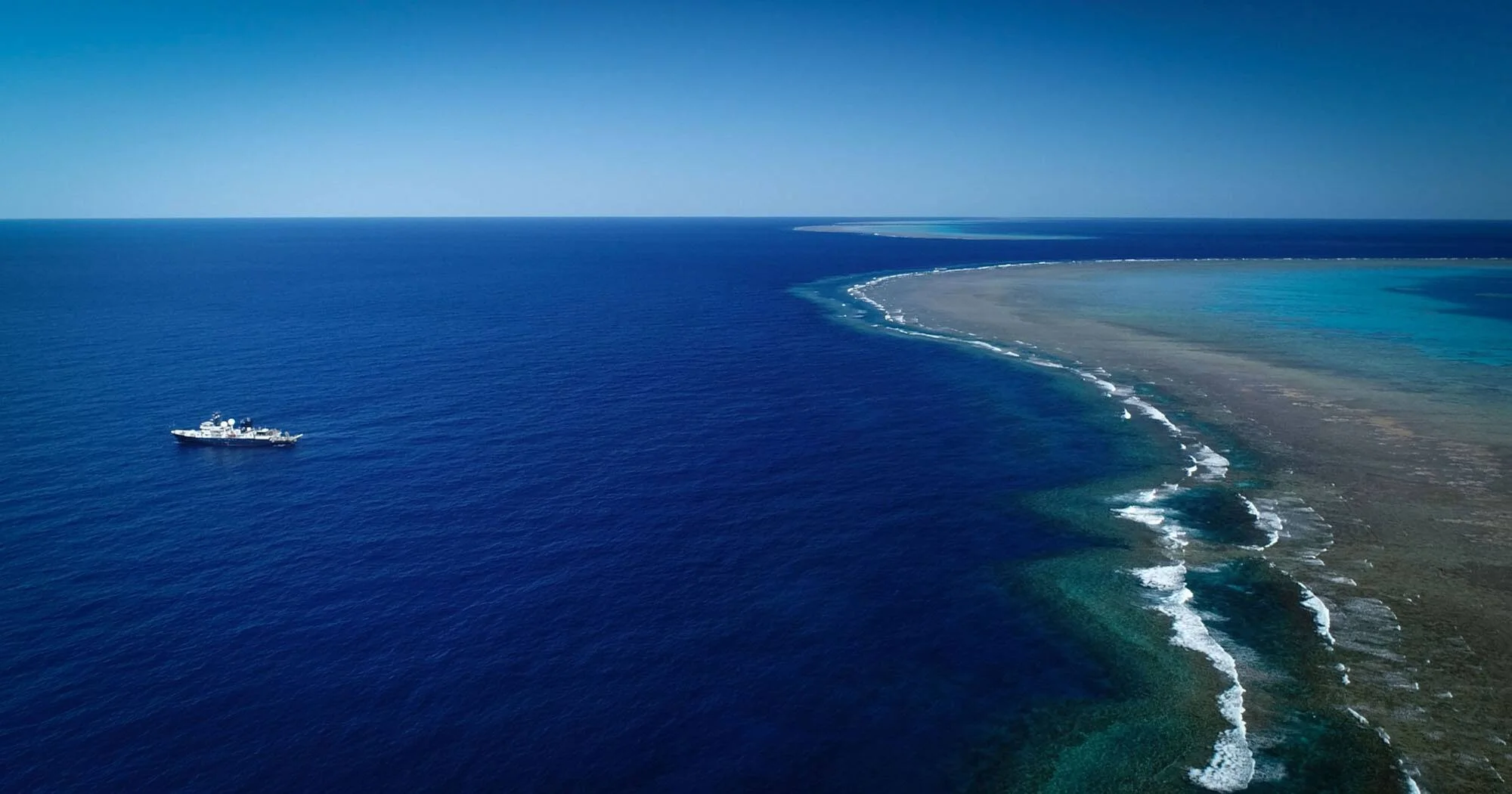 Vista aérea de un barco navegando en océano abierto junto al borde de un extenso arrecife de coral con aguas turquesas.