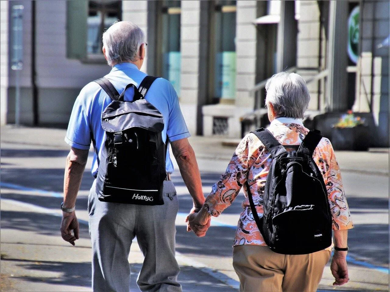 Pareja de personas mayores con mochilas negras camina tomada de la mano por una calle urbana soleada.