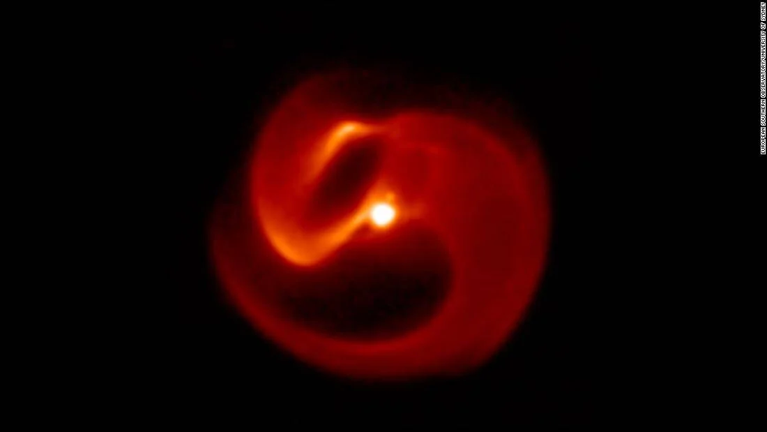 Imagen astronómica en tonos rojos que muestra espirales de gas y polvo alrededor de un sistema estelar binario con núcleo brillante central.