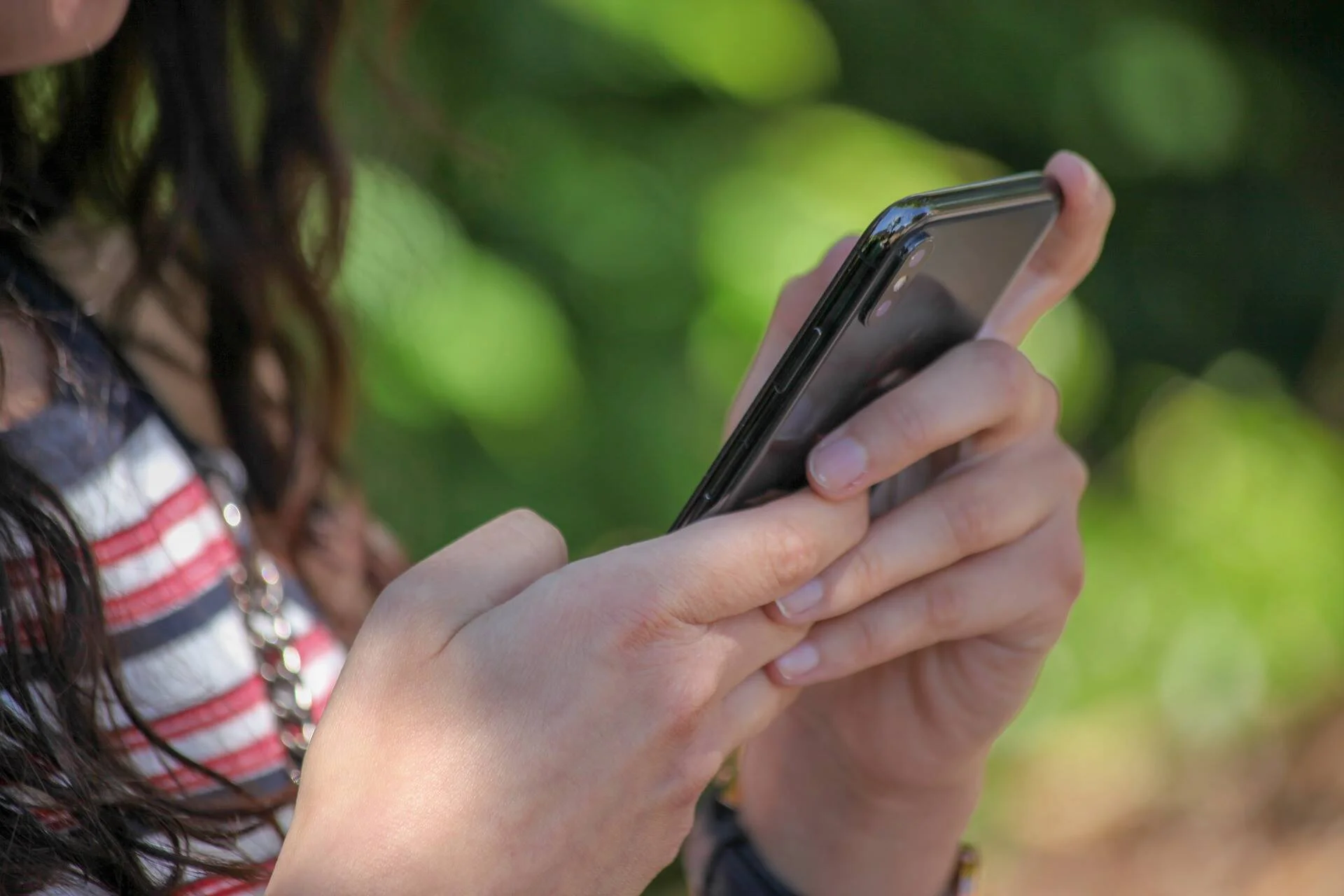 Manos de una joven sosteniendo y escribiendo en un teléfono inteligente con fondo verde desenfocado.