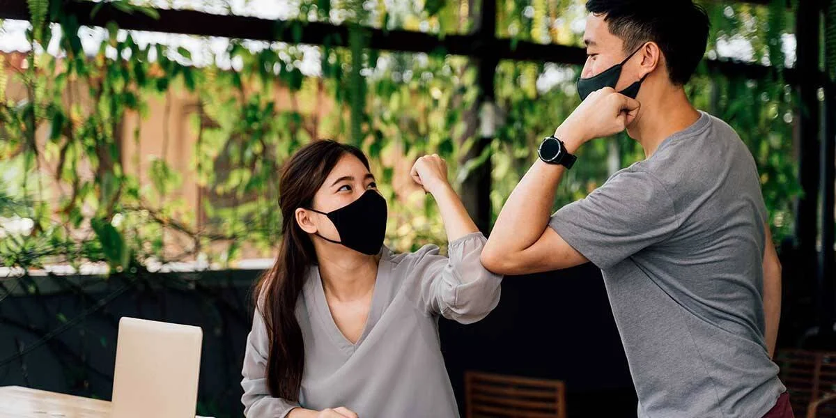 Dos personas con mascarillas negras se saludan chocando los puños como medida de distanciamiento social.
