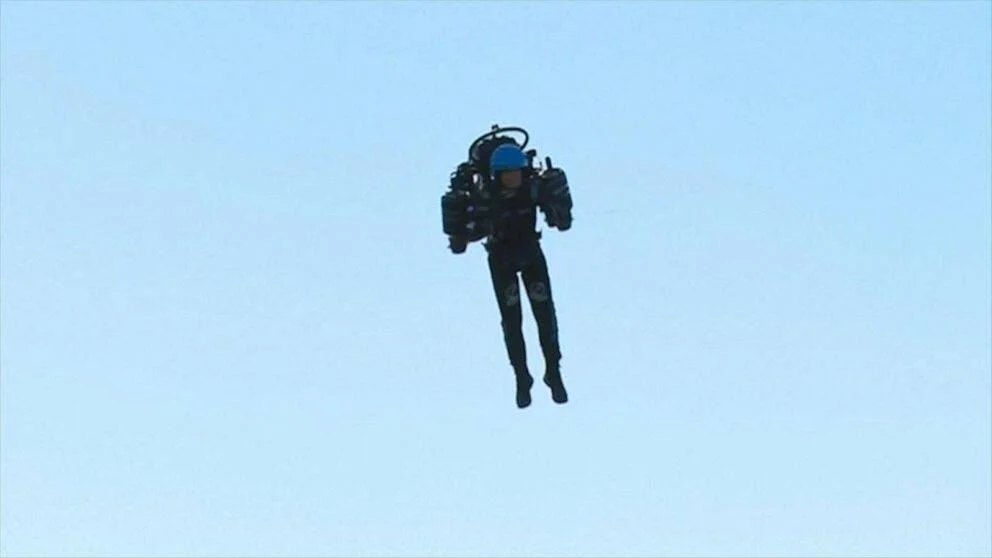 Una persona equipada con un jetpack vuela en el cielo despejado, llevando casco azul y traje negro.