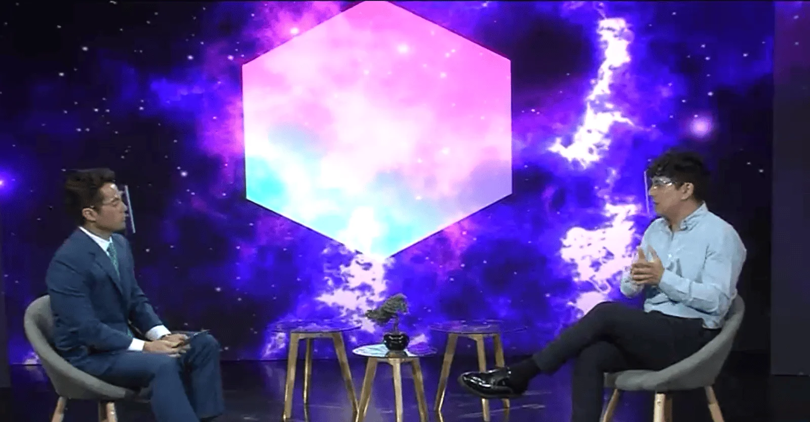 Dos hombres sentados en sillones frente a frente en un set de televisión con fondo galáctico púrpura y cubo luminoso rosa.