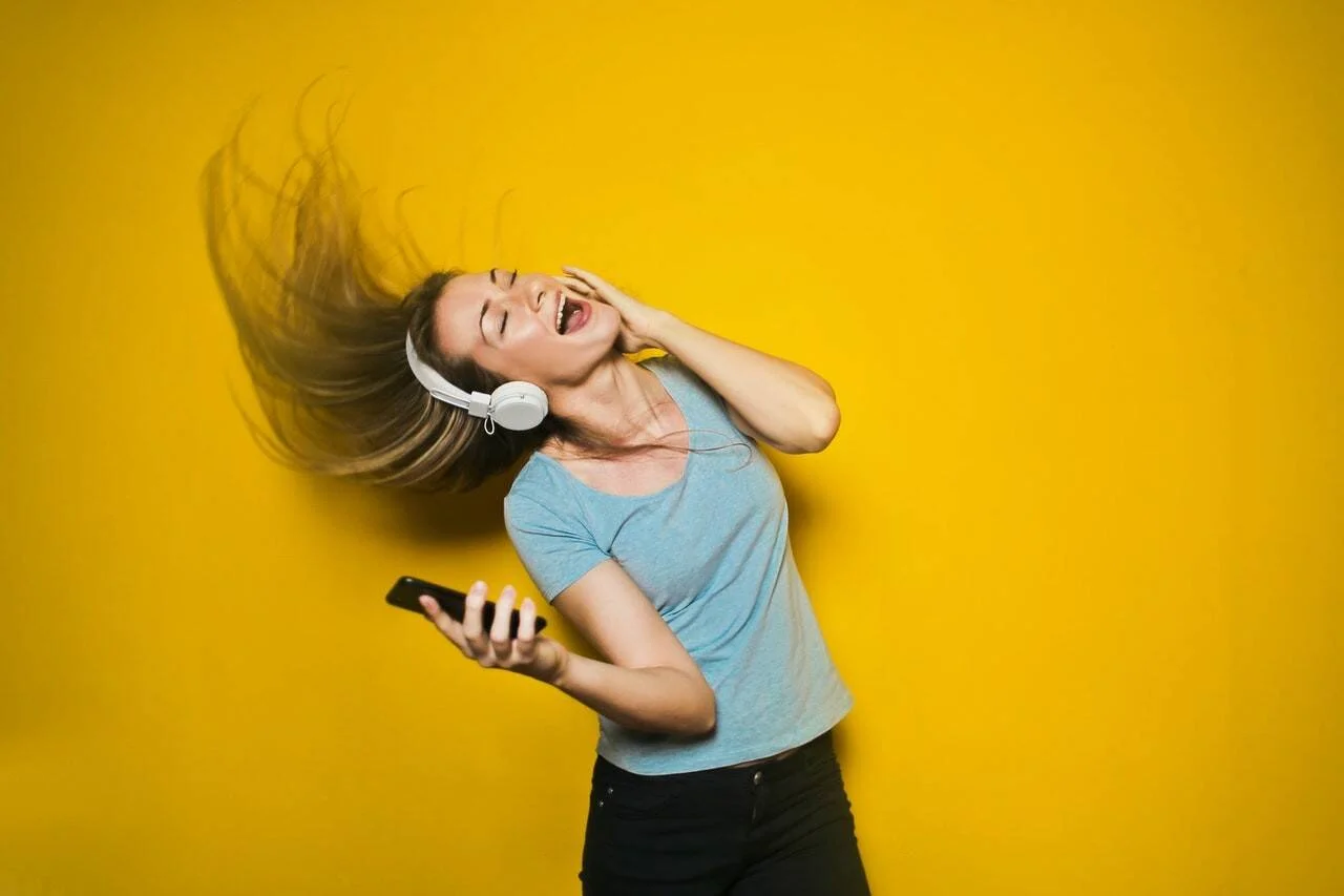 Mujer joven baila eufórica con auriculares blancos y teléfono en mano, agitando el cabello sobre fondo amarillo.