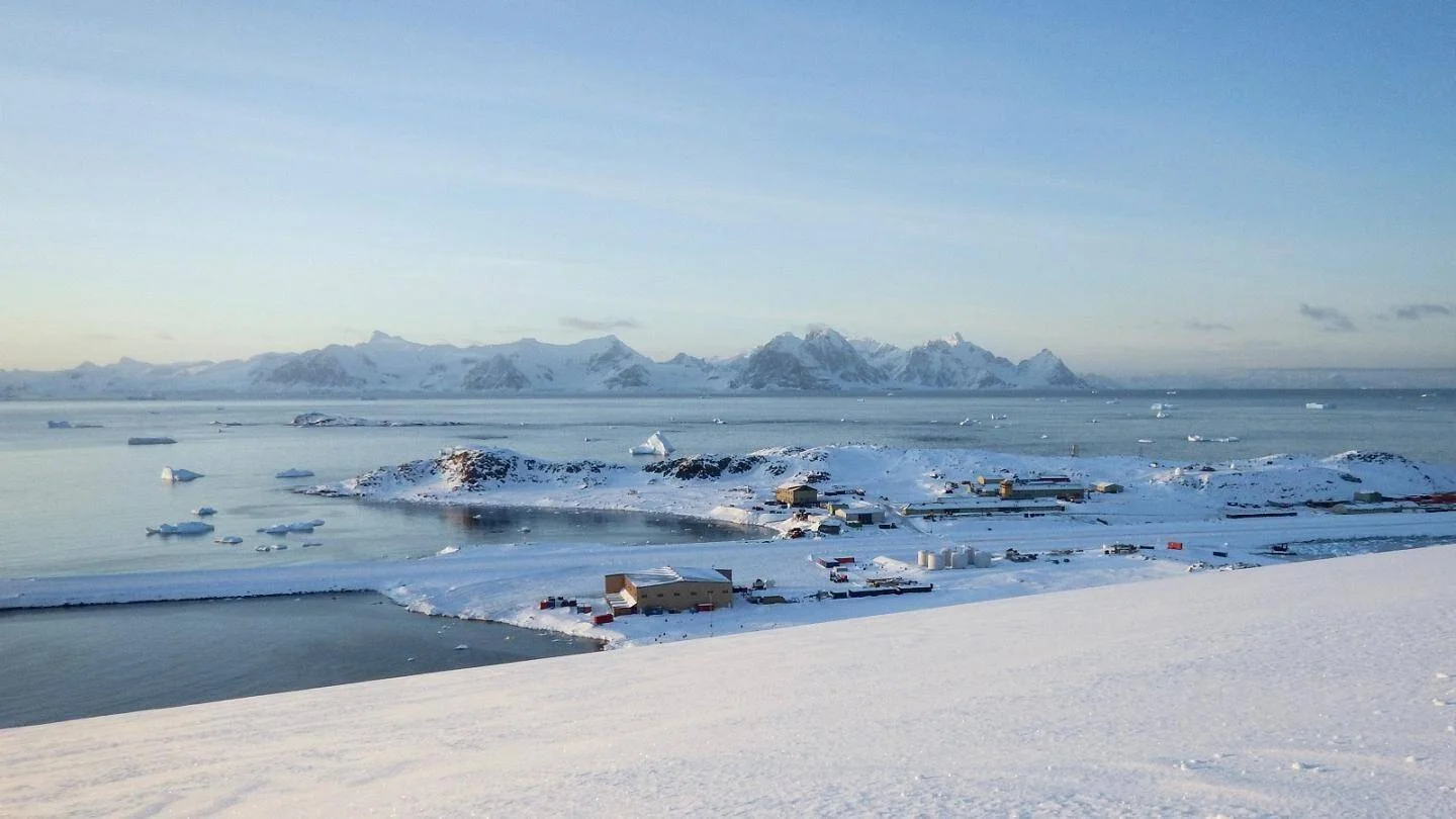 Vista aérea de una base científica antártica rodeada de nieve, con icebergs flotando en el mar y montañas nevadas al fondo.