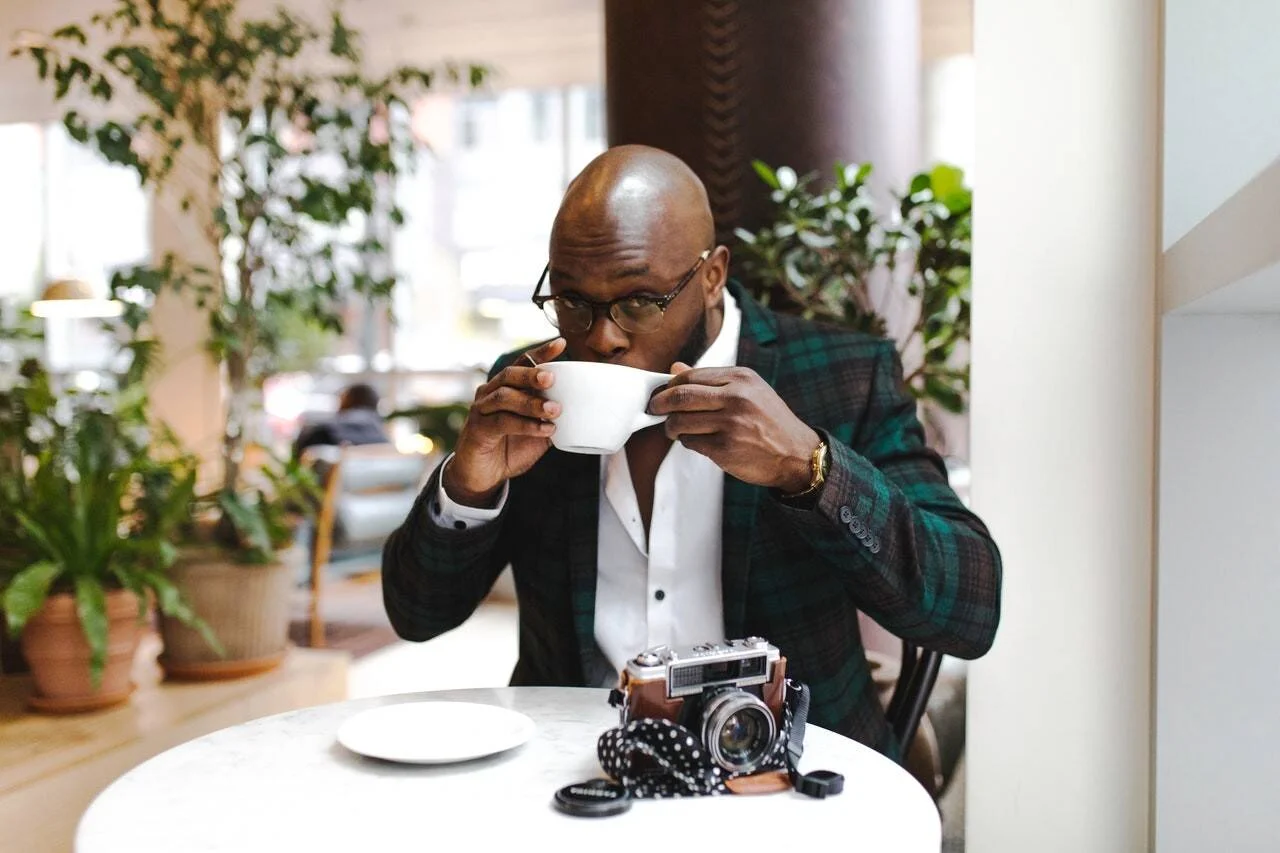 Hombre con blazer a cuadros y gafas bebe una taza de café en una mesa donde reposa una cámara fotográfica analógica.