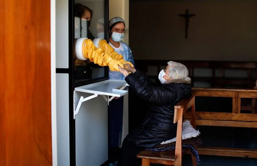 Una anciana con mascarilla recibe la comunión a través de una ventanilla de una sacerdotisa con equipo de protección en una iglesia.
