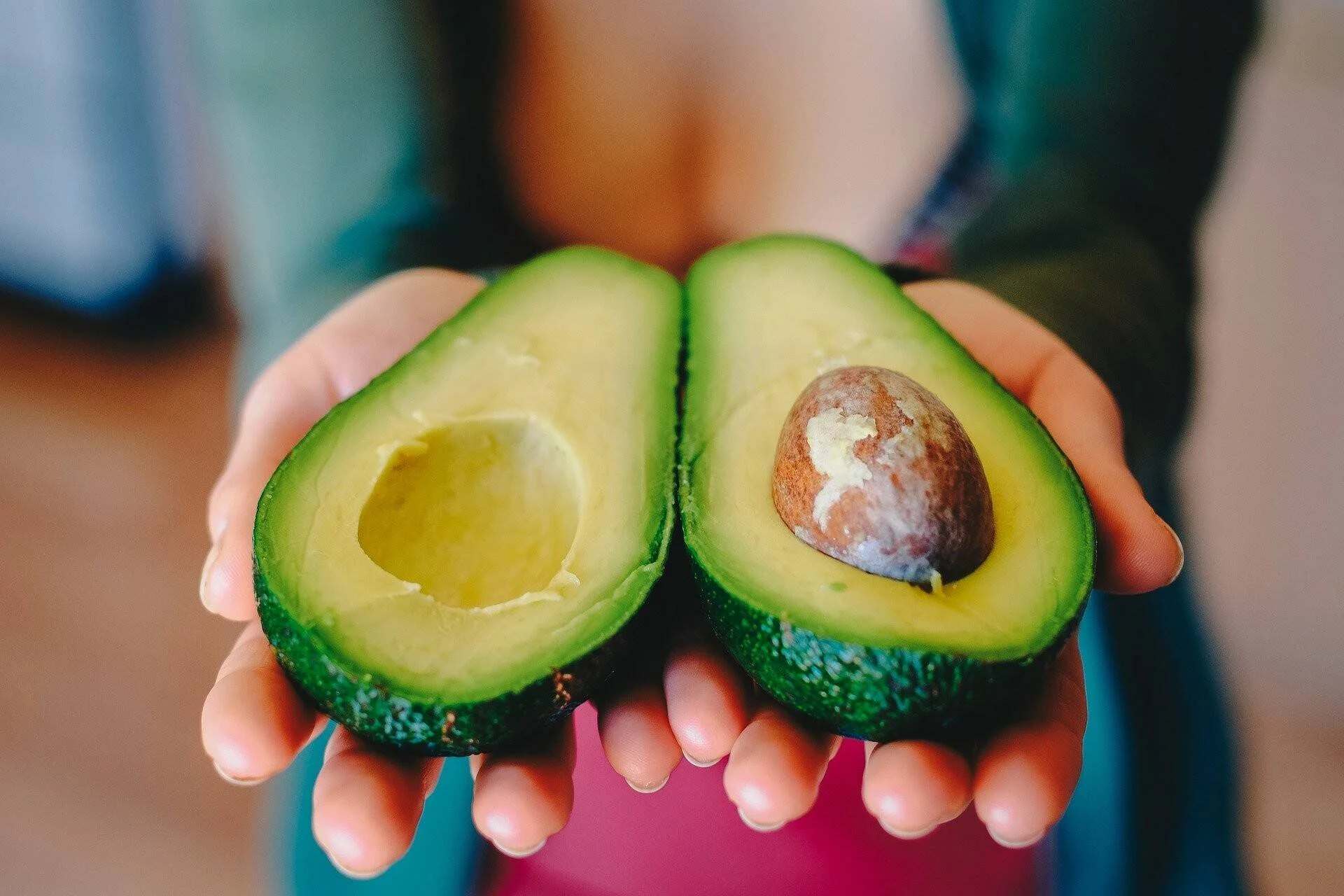Dos manos sostienen las mitades de un aguacate maduro abierto, mostrando su pulpa verde y el hueso marrón.