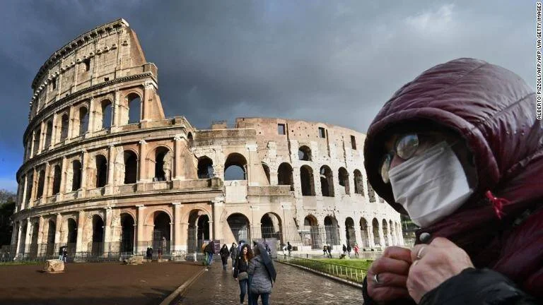Persona con mascarilla sanitaria y capucha frente al Coliseo de Roma bajo un cielo nublado.