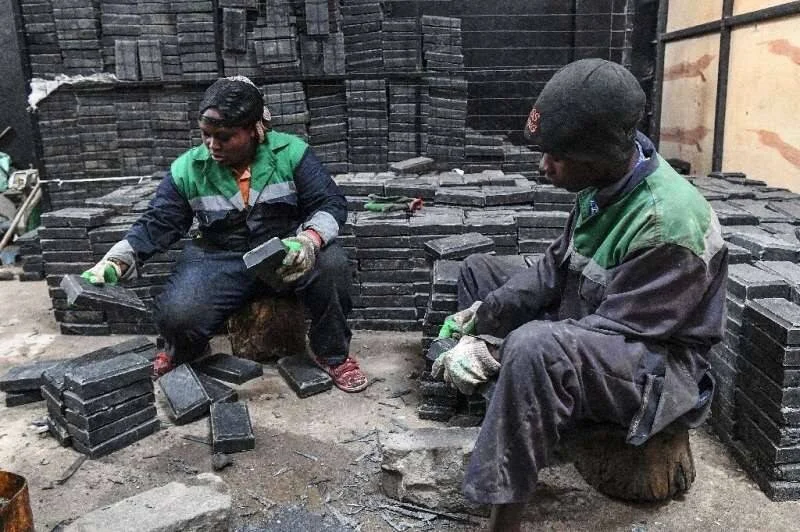 Dos trabajadores con uniformes verdes clasifican y apilan bloques rectangulares de carbón en un taller industrial.
