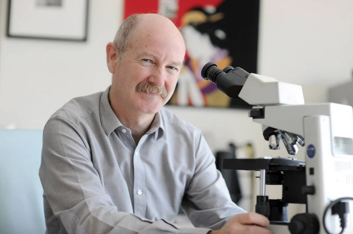 Hombre de mediana edad con bigote posa sonriente junto a un microscopio óptico en un entorno de laboratorio.