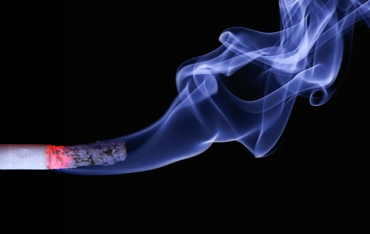 Cigarrillo encendido con brasa roja desprendiendo volutas de humo azul sobre fondo negro.