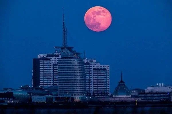 Gran luna llena de tonos rojizos se eleva sobre el horizonte de una ciudad con edificios iluminados en la noche.