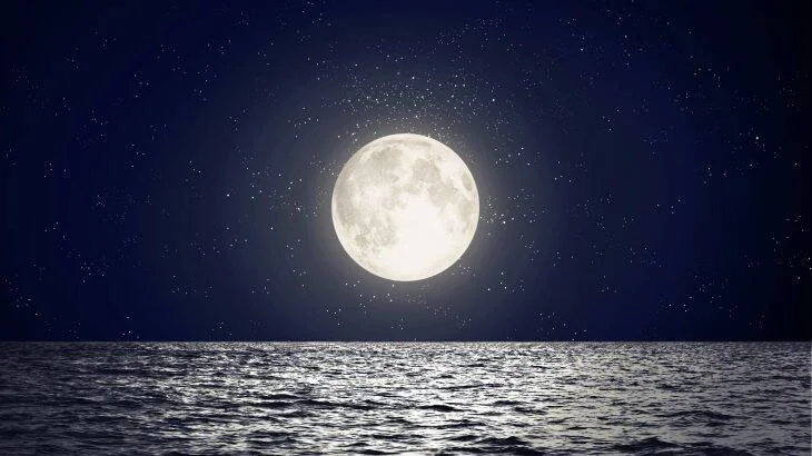 Luna llena y brillante se refleja sobre el mar en un cielo nocturno estrellado.