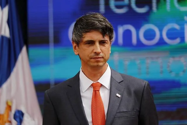 Hombre de traje gris oscuro y corbata naranja posando en un acto oficial con la bandera de Chile al fondo.